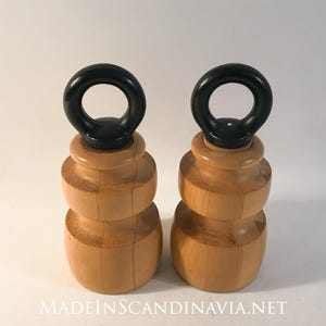 May include: Two wooden objects with a light brown finish and a black ring on top. The objects have a layered design and are set against a white background. The text "MADEINSCANDINAVIA.NET" is visible at the bottom.