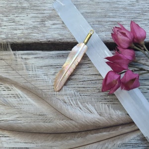 Puede incluir: Un colgante de pluma nacarada con una fianza dorada. El colgante está sobre una superficie de madera con un cristal blanco y flores rosas secas. Dos plumas más están en primer plano.