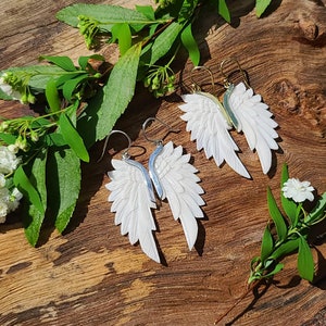 May include: A pair of white carved angel wing earrings with silver and gold hooks. The earrings are displayed on a wooden surface with green leaves and white flowers.