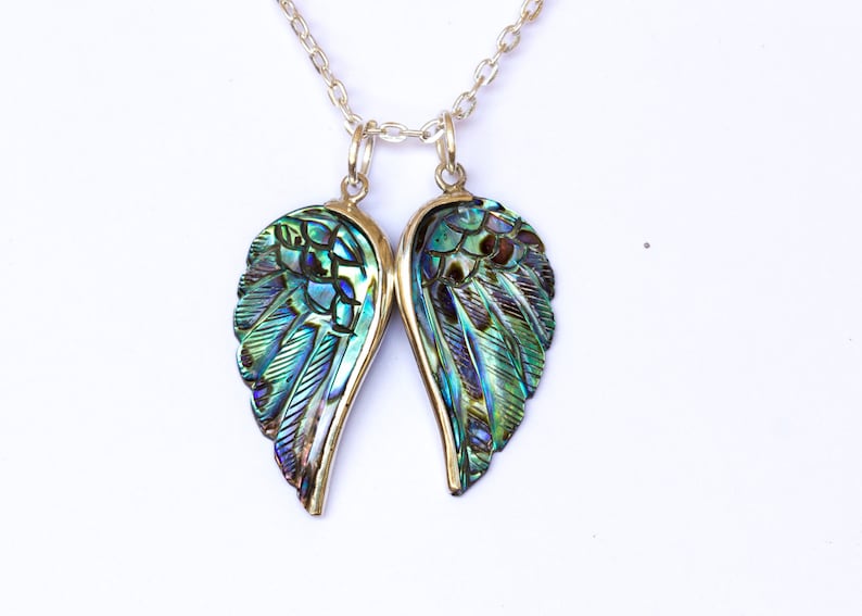 Puede incluir: Un collar de cadena de plata con un colgante de alas de &aacute;ngel de concha de abul&oacute;n iridiscente. Las alas est&aacute;n talladas con un patr&oacute;n de plumas y tienen un color verde, azul y morado.