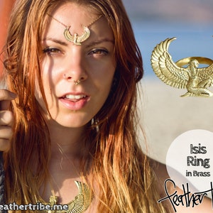 May include: A gold-toned ring featuring a detailed depiction of the Egyptian goddess Isis with outstretched wings. The ring is labeled "Isis Ring in Brass" and "Feather Tribe".