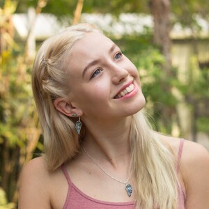 Puede incluir: Una joven con el pelo largo y rubio sonr&iacute;e y mira hacia arriba. Lleva un top rosa sin mangas y un collar plateado con un colgante azul y verde. Tambi&eacute;n lleva pendientes plateados con detalles azules y verdes.