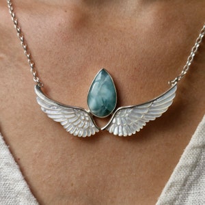 Könnte beinhalten: Eine silberne Halskette mit einem tropfenförmigen, hellblauen Edelsteinanhänger. Der Anhänger ist zwischen zwei weißen, flügelförmigen Elementen angeordnet. Die Halskette wird am Hals getragen.