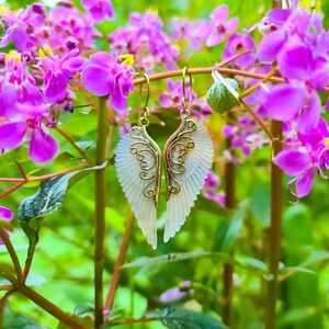 K&ouml;nnte beinhalten: Ein Paar goldene Engelsfl&uuml;gel-Ohrringe mit wei&szlig;en, schillernden Fl&uuml;geln. Die Ohrringe h&auml;ngen an einem Zweig mit rosa Bl&uuml;ten.