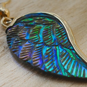 Puede incluir: Un pendiente de oro con un colgante en forma de ala iridiscente azul y verde. El colgante tiene una superficie texturizada y un borde dorado.