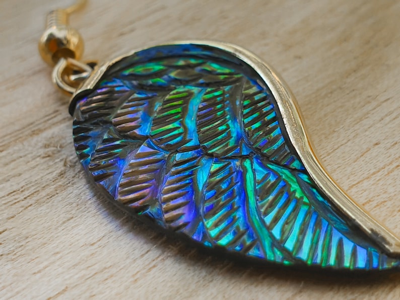 Puede incluir: Un pendiente de oro con un colgante en forma de ala iridiscente azul y verde. El colgante tiene una superficie texturizada y un borde dorado.