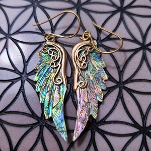 May include: A pair of gold-tone earrings with iridescent wings. The wings are made of a material that shimmers with a variety of colors, including green, blue, and purple.