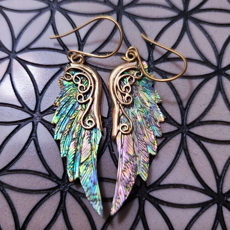 May include: A pair of gold-tone earrings with iridescent wings. The wings are made of a material that shimmers with a variety of colors, including green, blue, and purple.