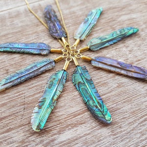 Puede incluir: Un conjunto de siete colgantes iridiscentes con forma de pluma dispuestos en un patrón circular. Cada colgante cuenta con una anilla metálica dorada y está sujeto a una fina cadena de color oro, mostrando tonos brillantes en azul, verde y morado.