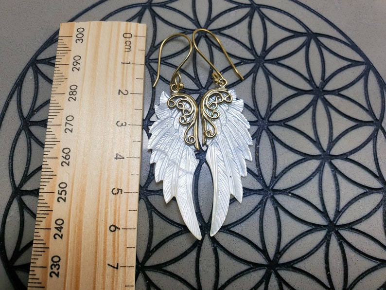 May include: A pair of gold and white angel wing earrings. The wings are made of mother of pearl and have a delicate filigree design. The earrings are approximately 6 cm long.