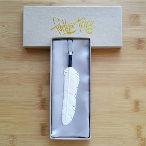 Puede incluir: Un colgante de pluma blanca tallada con un cordón negro, presentado en una caja rectangular. La caja es de color marrón claro con el texto dorado "feather tribe". El colgante se muestra sobre una tela satinada.
