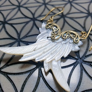 May include: Gold and white shell earrings with a delicate, ornate design. The earrings feature a pair of wings made from white shell with a gold filigree design.