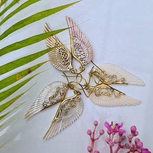 K&ouml;nnte beinhalten: Ein Paar goldene und irisierende wei&szlig;e Engelsfl&uuml;gel-Ohrringe. Die Fl&uuml;gel sind aus filigranem Metall gefertigt und haben eine perlmuttartige Oberfl&auml;che.