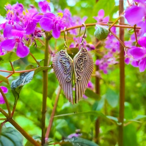 K&ouml;nnte beinhalten: Ein Paar goldene Engelsfl&uuml;gel-Ohrringe mit filigranen Details. Die Fl&uuml;gel sind hellrosa und haben ein zartes, feminines Design.