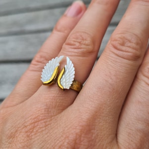 May include: A gold ring with white wings. The wings are made of a pearlescent material and are shaped like angel wings.