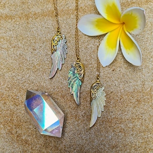 K&ouml;nnte beinhalten: Drei goldene Halsketten mit schillernden Fl&uuml;gelanh&auml;ngern. Die Fl&uuml;gel sind aus Perlmutt gefertigt und haben ein zartes, verziertes Design. Die Halsketten werden auf einer sandigen Oberfl&auml;che mit einer wei&szlig;en Blume pr&auml;sentiert.