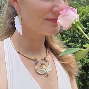 May include: A gold-toned necklace with a pendant featuring a scarab beetle design, adorned with white enamel and blue gemstones. Matching white wing-shaped earrings with gold accents are also visible. The jewelry is worn with a white top.