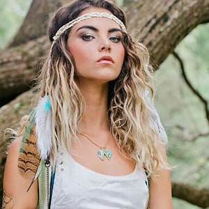 Puede incluir: Una mujer con el pelo largo y ondulado rubio que lleva una diadema blanca con un dise&ntilde;o trenzado. Lleva un top blanco sin mangas y un collar de oro con un colgante turquesa. Lleva una pluma atada al pelo.