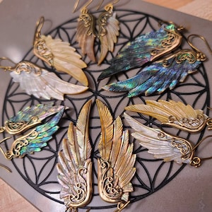 May include: A set of six pairs of earrings featuring iridescent abalone shell wings with gold accents. The wings are shaped like angel wings and have intricate details.
