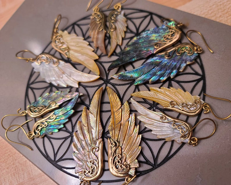 May include: A set of six pairs of earrings featuring iridescent abalone shell wings with gold accents. The wings are shaped like angel wings and have intricate details.