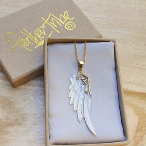 Puede incluir: Un collar de cadena dorada con un colgante de ala de ángel de nácar blanco. El colgante tiene un diseño de filigrana dorada y está en una pequeña caja de regalo.