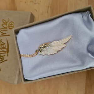 K&ouml;nnte beinhalten: Eine goldene Halskette mit einem wei&szlig;en und goldenen Engelsfl&uuml;gel-Anh&auml;nger. Der Anh&auml;nger befindet sich in einer kleinen Schachtel mit der Aufschrift "Feather Tribe" auf der Seite.