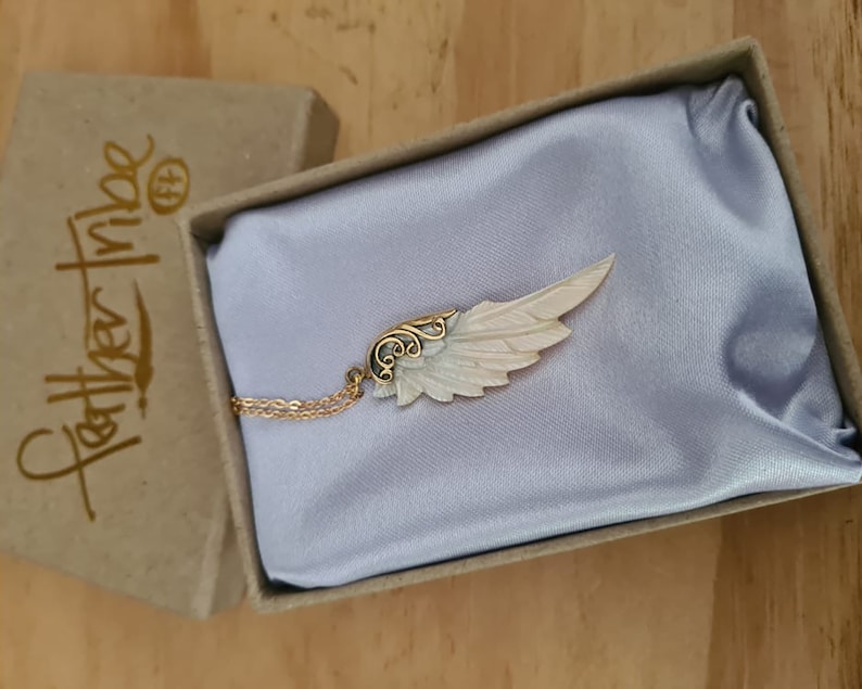 K&ouml;nnte beinhalten: Eine goldene Halskette mit einem wei&szlig;en und goldenen Engelsfl&uuml;gel-Anh&auml;nger. Der Anh&auml;nger befindet sich in einer kleinen Schachtel mit der Aufschrift "Feather Tribe" auf der Seite.