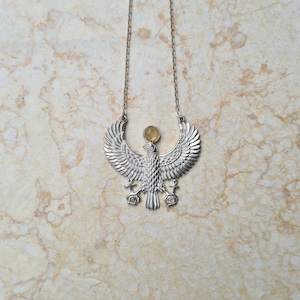 Puede incluir: Collar de plata con un colgante de pájaro alado detallado y una piedra preciosa amarilla. El colgante está suspendido de una cadena de plata.