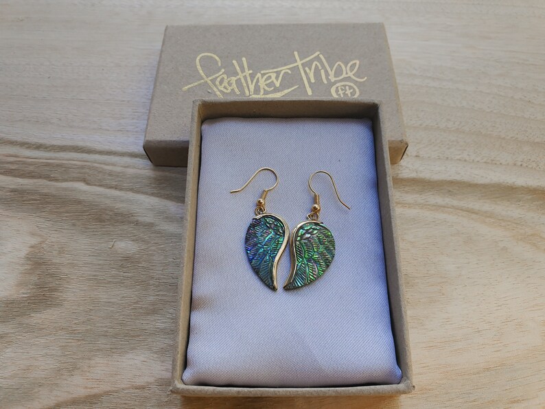 Puede incluir: Un par de pendientes de oro con alas iridiscentes verdes y azules. Los pendientes est&aacute;n en una peque&ntilde;a caja con las palabras "Feather Tribe" escritas en ella.
