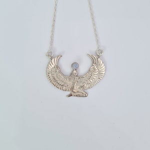 Puede incluir: Collar de plata con un colgante detallado de la diosa egipcia Isis con alas extendidas y una piedra de luna en el centro.