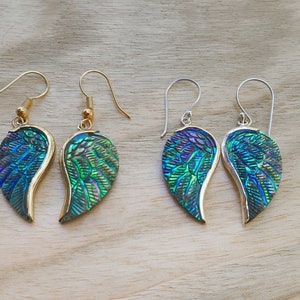 Puede incluir: Un par de pendientes en forma de ala de color azul y verde iridiscente con ganchos de oro y plata.