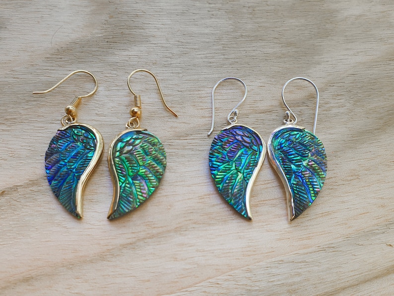 Puede incluir: Un par de pendientes en forma de ala de color azul y verde iridiscente con ganchos de oro y plata.