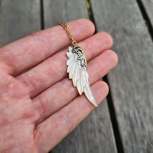 K&ouml;nnte beinhalten: Eine goldene Kettenkette mit einem wei&szlig;en und goldenen Engelsfl&uuml;gel-Anh&auml;nger. Der Fl&uuml;gel ist mit filigranen Schnitzereien verziert und hat eine zarte, feminine Ausstrahlung.