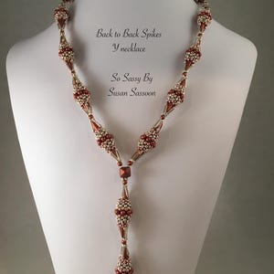 Könnte beinhalten: Eine Y-förmige Halskette mit einem Perlendesign in Braun- und Goldtönen. Die Halskette weist eine Reihe von stachelartigen Formationen auf und wird auf einer weißen Schaufensterpuppe präsentiert. Der Text "Back to Back Spikes Y necklace" und "So Sassy By Susan Sassoon" ist sichtbar.