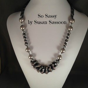 Peut inclure: Un collier de perles grises et de petites perles noires et transparentes. Le collier est présenté sur un mannequin blanc et porte le texte "So Sassy by Susan Sassoon".