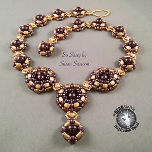 Op de afbeelding: Een gouden en paarse kralenketting met een hanger. De ketting is gemaakt van kleine, ronde kralen en heeft een geometrisch ontwerp. De hanger is vierkant met een ronde kraal in het midden. De ketting heet "So Sassy" van Susan Sassoon.