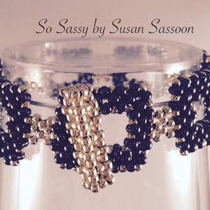 May include: A beaded bracelet with a geometric design, featuring navy blue and silver beads. The bracelet is displayed on a clear glass. The text "So Sassy by Susan Sassoon" is above the bracelet.