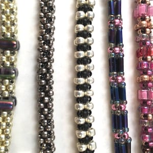 Könnte beinhalten: Fünf Perlenarmbänder in verschiedenen Farben und Mustern. Die Armbänder sind aus kleinen Perlen in verschiedenen Farben gefertigt, darunter Weiß, Schwarz, Silber, Rosa und Lila.