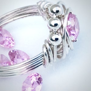 May include: A silver wire wrapped ring with a pink gemstone and three silver beads. The ring is surrounded by loose pink gemstones.