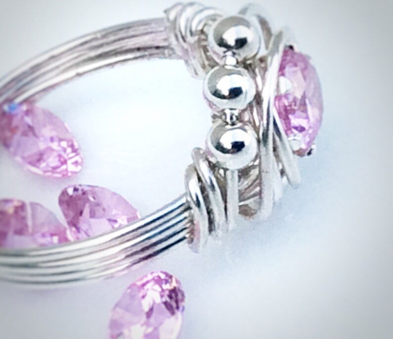 May include: A silver wire wrapped ring with a pink gemstone and three silver beads. The ring is surrounded by loose pink gemstones.