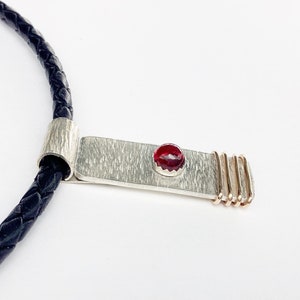 May include: A silver pendant necklace with a red gemstone and gold accents hangs from a black leather cord.