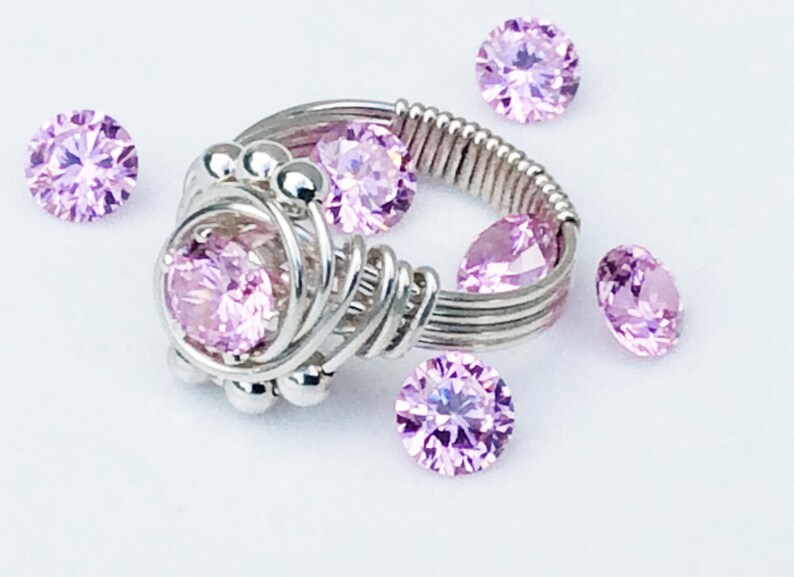 May include: A silver wire wrapped ring with a large pink gemstone in the center. The ring is surrounded by smaller pink gemstones.
