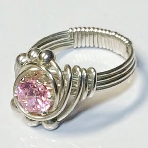 May include: A silver wire wrapped ring with a pink gemstone in the center. The ring is made with multiple strands of silver wire that are twisted together to create a unique design.