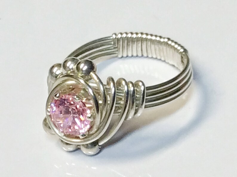 May include: A silver wire wrapped ring with a pink gemstone in the center. The ring is made with multiple strands of silver wire that are twisted together to create a unique design.
