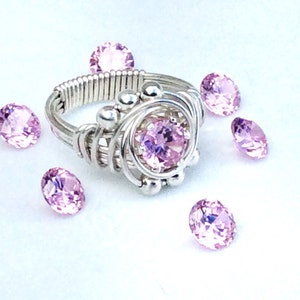 May include: A silver wire wrapped ring with a pink gemstone in the center. The ring is surrounded by small silver beads. There are several loose pink gemstones scattered around the ring.