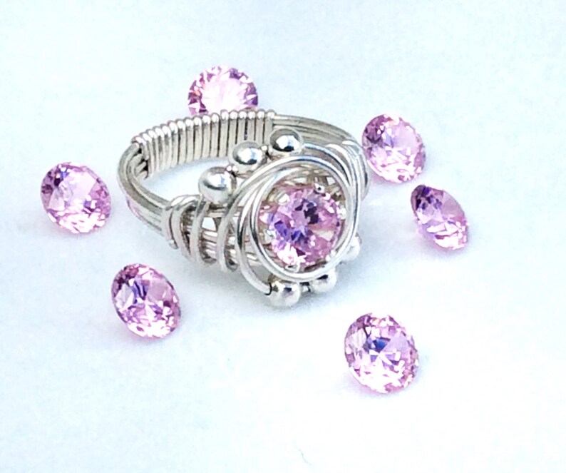 May include: A silver wire wrapped ring with a pink gemstone in the center. The ring is surrounded by small silver beads. There are several loose pink gemstones scattered around the ring.