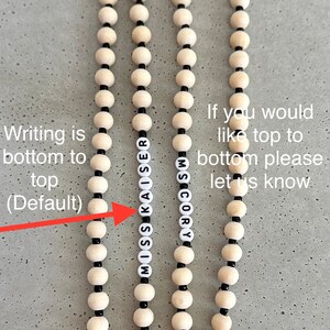 Może przedstawiać: Zbliżenie na dwa naszyjniki z drewnianych koralik&oacute;w z czarnymi koralikami. Naszyjniki tworzą napisy "MISS KAISER" i "SCORY". Tekst "Writing is bottom to top (Default)" jest napisany po lewej stronie obrazu, a tekst "If you would like top to bottom please let us know" jest napisany po prawej stronie obrazu.