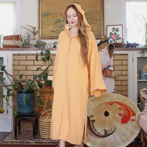 Op de afbeelding: Een lange, perzik kleurige gehoodde kaftan met lange mouwen. De kaftan heeft een opening in het midden met decoratieve stiksels en zijsplitten. De persoon draagt roze schoenen. Een strohoed ligt op de grond.