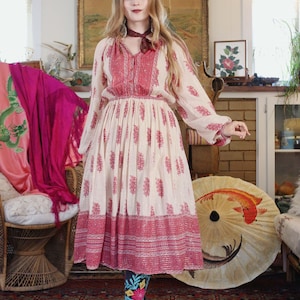 Könnte beinhalten: Cremefarbenes Kleid mit rotem Blumenmuster und langen Ärmeln. Das Kleid hat einen V-Ausschnitt, eine betonte Taille und einen fließenden Rock. Das Model trägt Stiefel mit Blumenmuster und einen bordeauxfarbenen Schal.