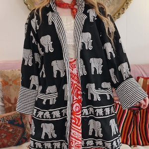 Puede incluir: Chaqueta negra estilo kimono con estampado de elefantes blancos y detalles a rayas. La chaqueta tiene cuello chal, mangas largas y bolsillos. La modelo lleva una falda estampada en rojo y blanco y una bufanda roja.